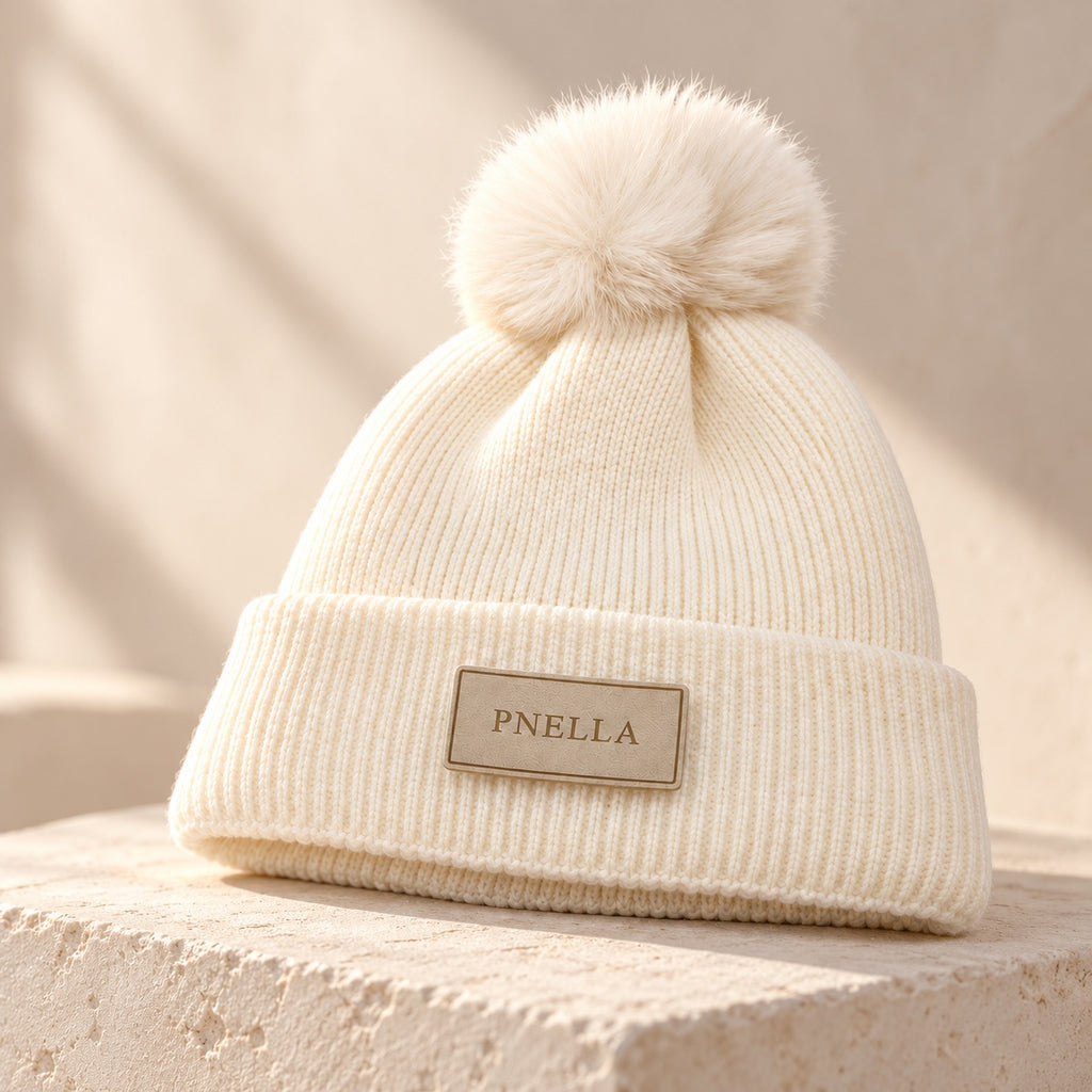 PNELLA cream pom-pom beanie styled on a stone plinth